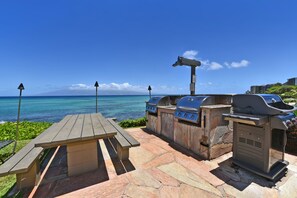 Eine Terrasse im Freien mit einem Holztisch und Grillanlagen mit Blick auf einen Strand.