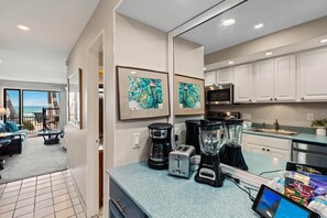 A modern kitchen with a coffee maker, toaster, blender, and a tablet on the counter. There are framed pictures on the wall and a view of the ocean through the sliding glass door. - Ocean views and A/C combine in this Deluxe studio (Lahaina)