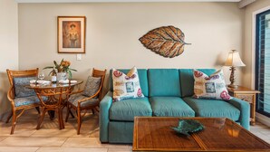 A living room with a teal sofa, wicker chairs, a coffee table, and a wall art piece. - Bright, Spacious Studio with Stunning Ocean Views (Lahaina)