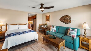 A hotel room with a bed, a teal sofa, a wooden coffee table, a ceiling fan, and a dining area with a table and chairs. - Bright, Spacious Studio with Stunning Ocean Views (Lahaina)