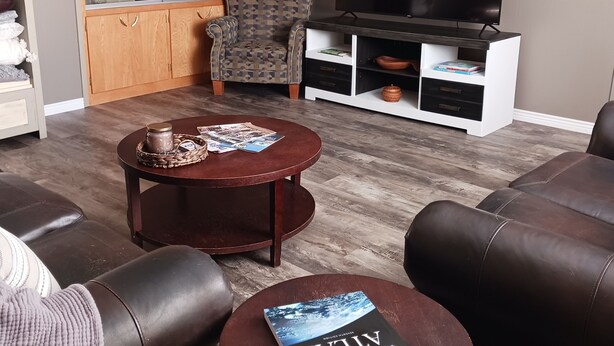 A living room with a brown leather sofa, a round coffee table with magazines, a patterned armchair, a wooden cabinet with glass doors, and a flat-screen TV on a white stand. - The Shopkeeper's Inn. 
Unique property with history, In the heart of Ely, Nv. (Ely)