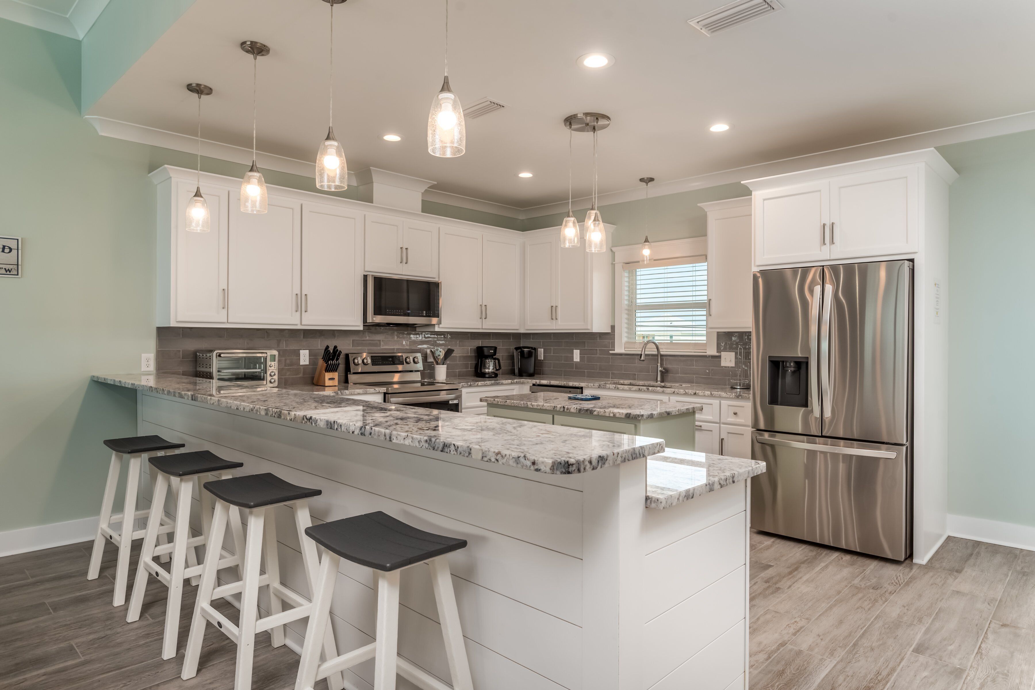 A modern kitchen with white cabinets, a stainless steel refrigerator, a marble island with bar stools, and pendant lighting.