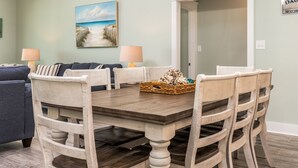 Un coin repas avec une table en bois, des chaises blanches et un tableau représentant une scène de plage accroché au mur.