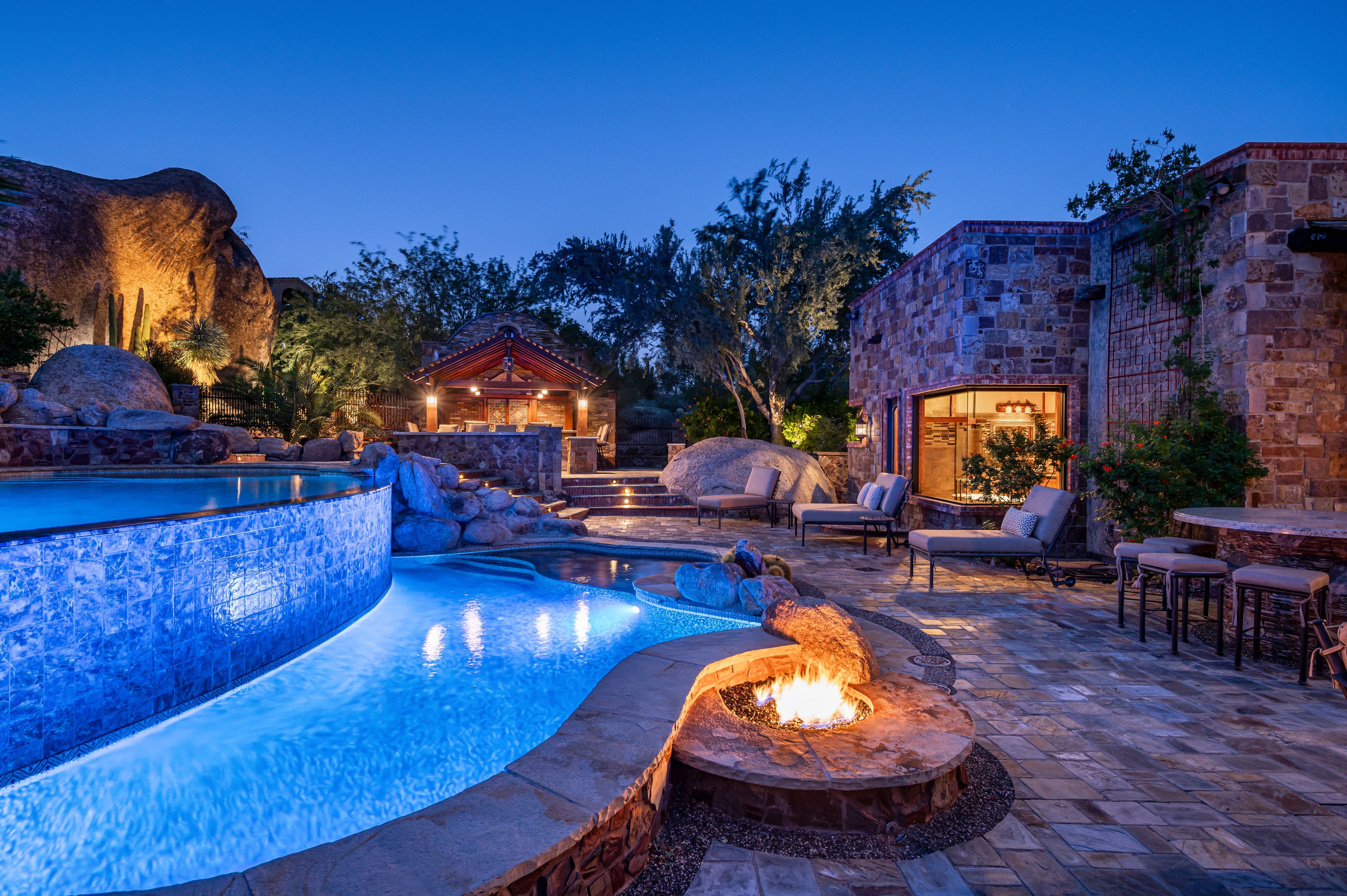 Zona de piscina con un hogar, sillas de descanso y un edificio de piedra con puerta de vidrio.