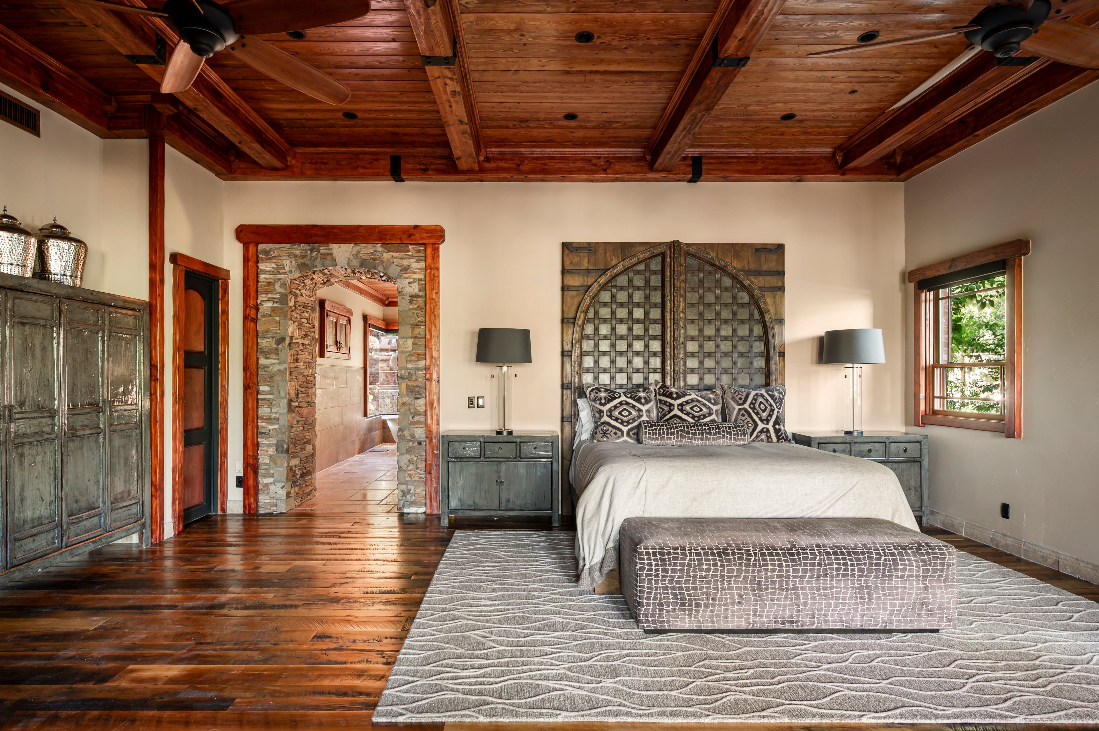 A bedroom with a wooden ceiling, a stone archway, a bed with patterned pillows, and a nightstand with a lamp.