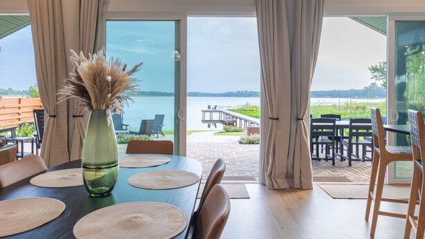 Un comedor con una mesa dispuesta para cuatro, un jarrón con flores secas y vistas a un muelle y follaje exterior.