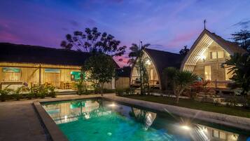 A pool area with a clear pool, wooden buildings, and a sunset sky.