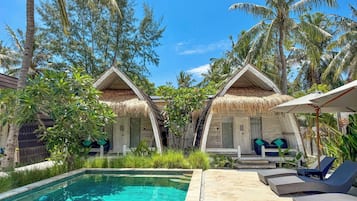A poolside area with two thatched-roof cottages, lounge chairs, and a clear pool.