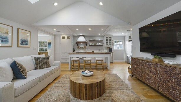 A spacious living area with a high ceiling, a central island with stools, a sofa with patterned cushions, a round wooden coffee table, and a TV on a cabinet. - Shell Mates - Luxury Lakefront Vacation Rental with Private Pool Space & Outdoor Area (Miramar Beach)