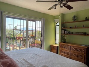 A bedroom with a bed, a ceiling fan, a dresser, and a window with a view of a balcony and outdoor area.