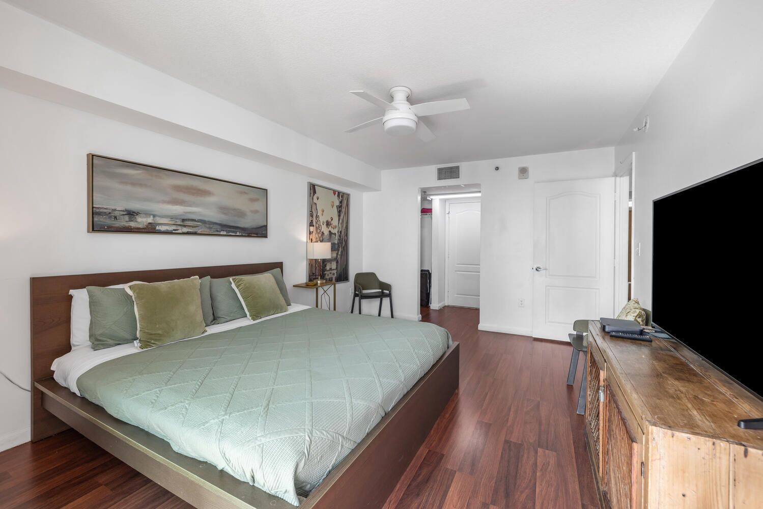 A bedroom with a bed, a desk with a TV, a chair, and a door leading to another room.