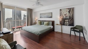 Een moderne hotelkamer met een groot bed, een bureau met een laptop, een stoel, een balkon met uitzicht op de stad, en een plafondventilator.