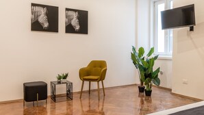 A room with a wooden floor, a yellow armchair, a black ottoman, two black and white zebra paintings, a large green plant, and a flat-screen TV mounted on the wall.
