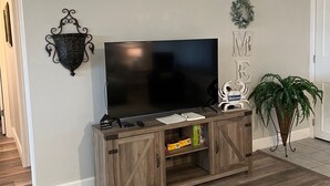 A living room with a TV on a wooden stand, a wall-mounted light fixture, a welcome sign, and a potted plant.
