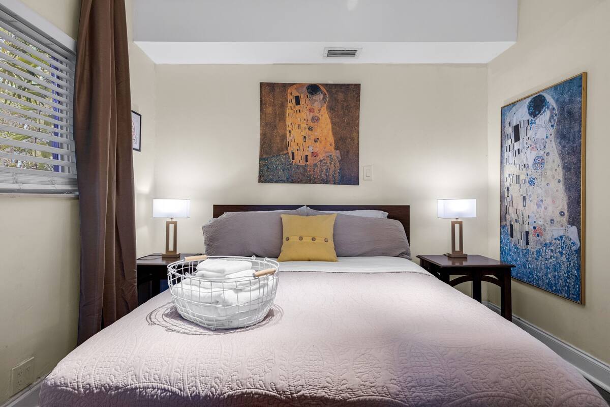 A hotel room with a large bed, two bedside tables, a wall-mounted painting, and a window with blinds.