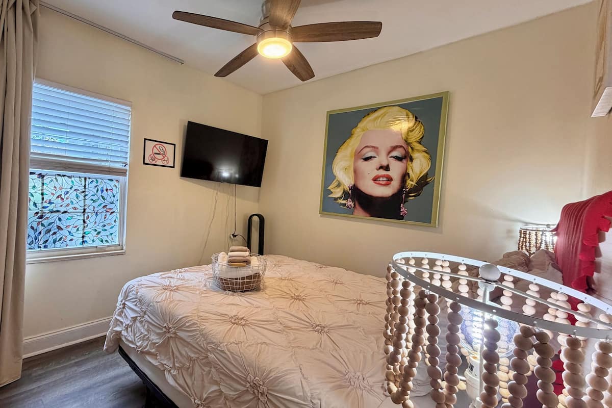 A bedroom with a bed, a ceiling fan, a TV, a window with blinds, and a painting.