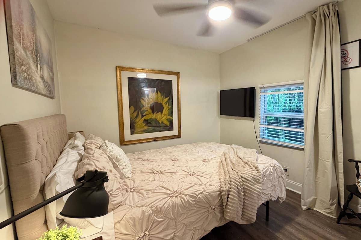 A bedroom with a bed, a ceiling fan, a TV, a window with blinds, a chair, and a framed picture on the wall.