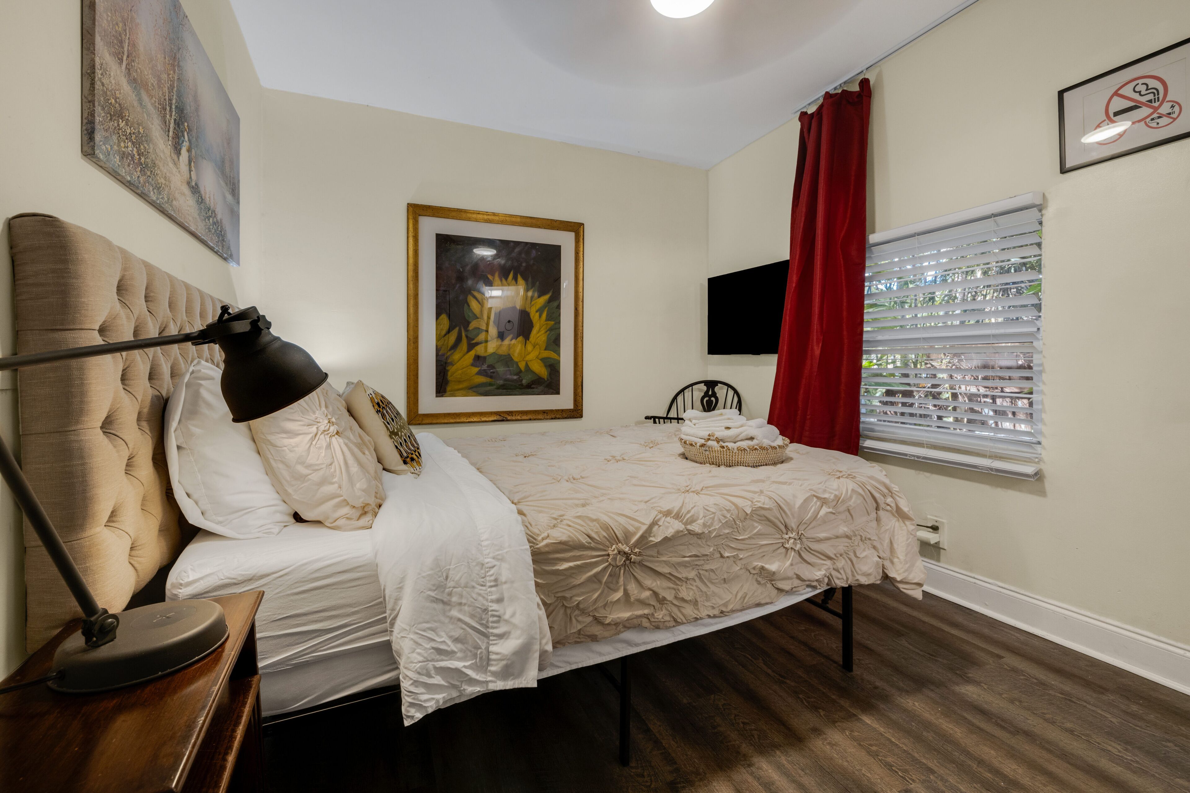 Um quarto com uma cama, luminária de cabeceira, televisão, janela com persianas e um quadro na parede.