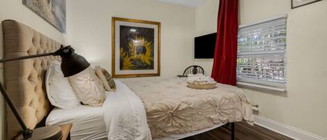 A bedroom with a bed, a bedside lamp, a television, a window with blinds, and a painting on the wall.
