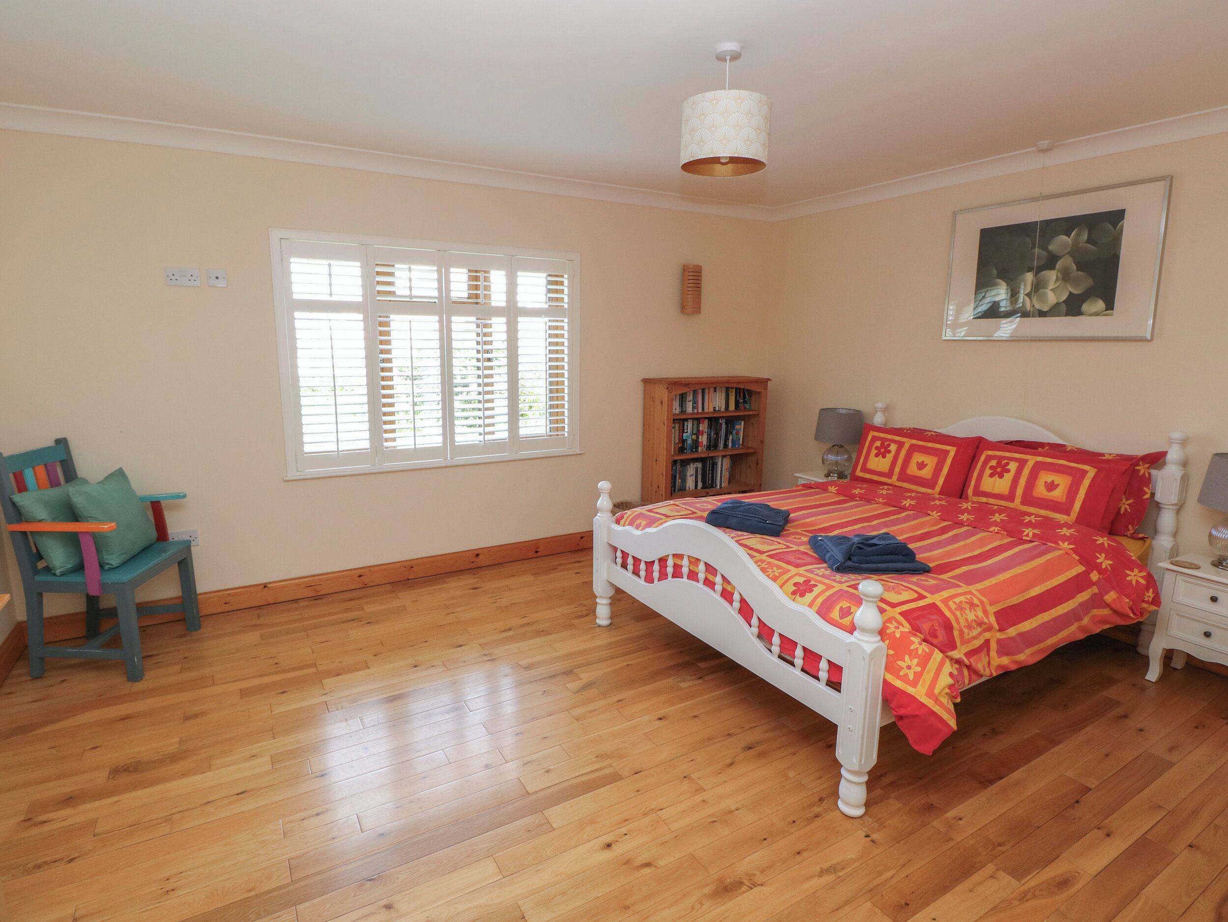 A bedroom with a wooden floor, a bed with a red and yellow patterned bedspread, a white headboard, a wooden bookshelf, a chair with colorful cushions, a window with blinds, and a ceiling light fixture.