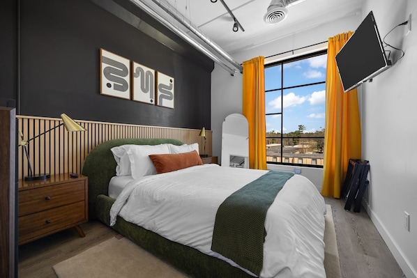A modern bedroom with a bed, a nightstand, a desk, a chair, a window with curtains, and a mounted TV.