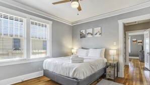 Une chambre moderne avec un lit gris, du linge de lit blanc, un plancher en bois et un ventilateur de plafond.