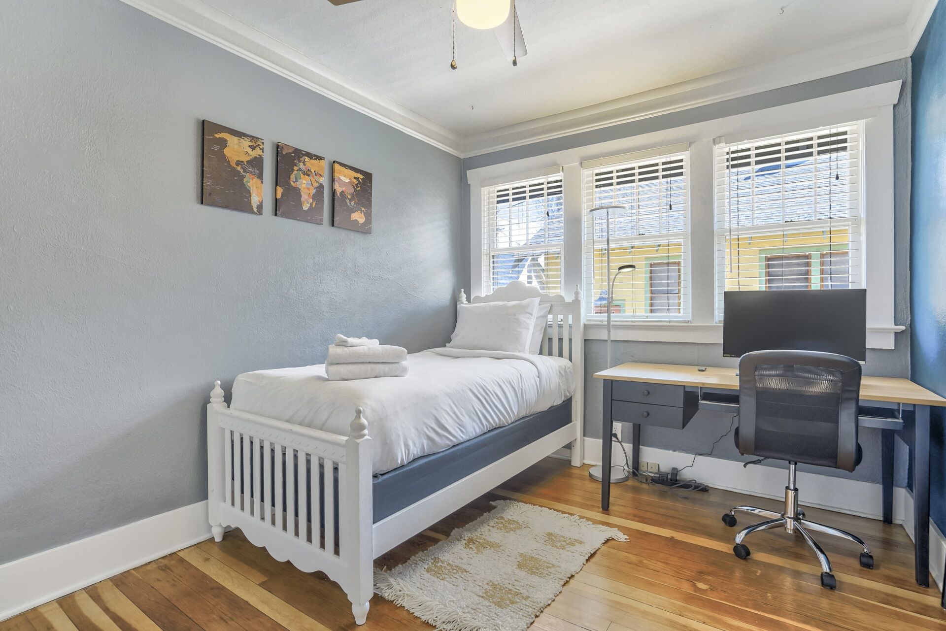 Ein Schlafzimmer mit einem Bett, einem Schreibtisch und einem Stuhl. Drei Bilder hängen an der Wand und ein Fenster mit Jalousien ist vorhanden.