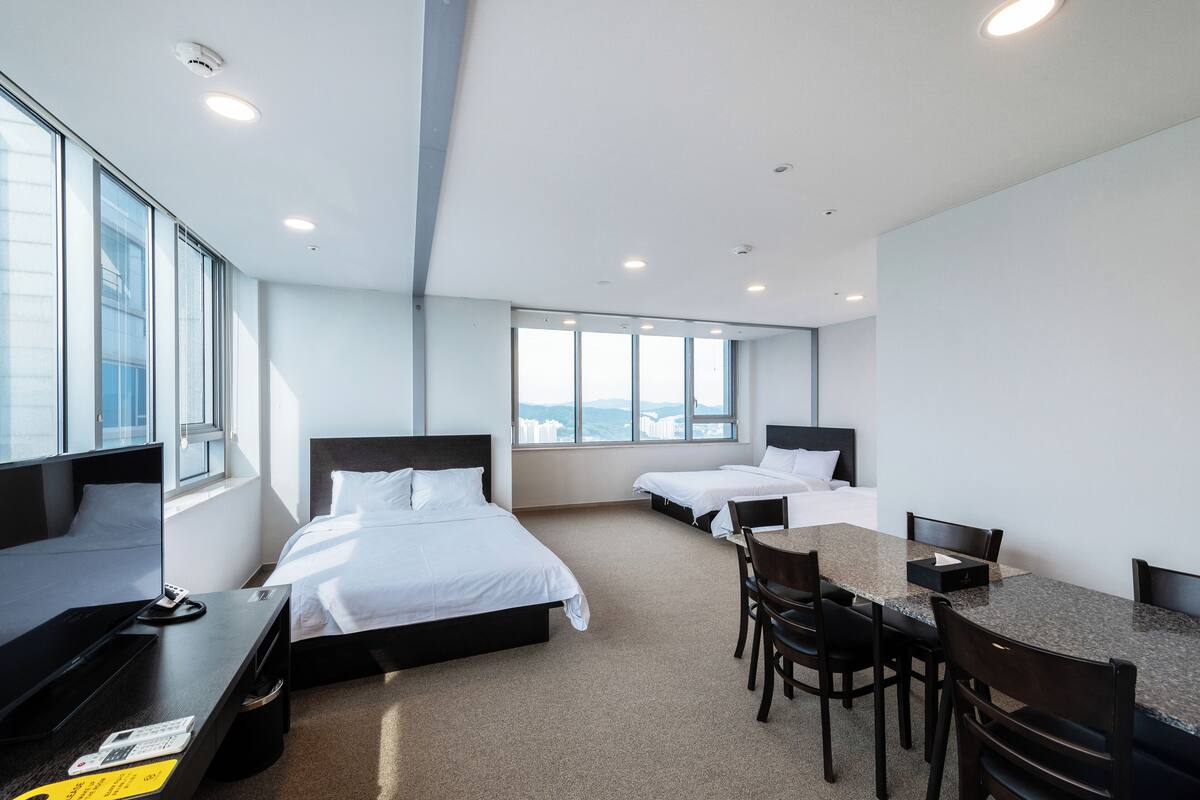 Une chambre d’hôtel avec deux lits simples, un bureau avec une chaise, une table à manger avec des chaises et de grandes fenêtres donnant sur la ville.