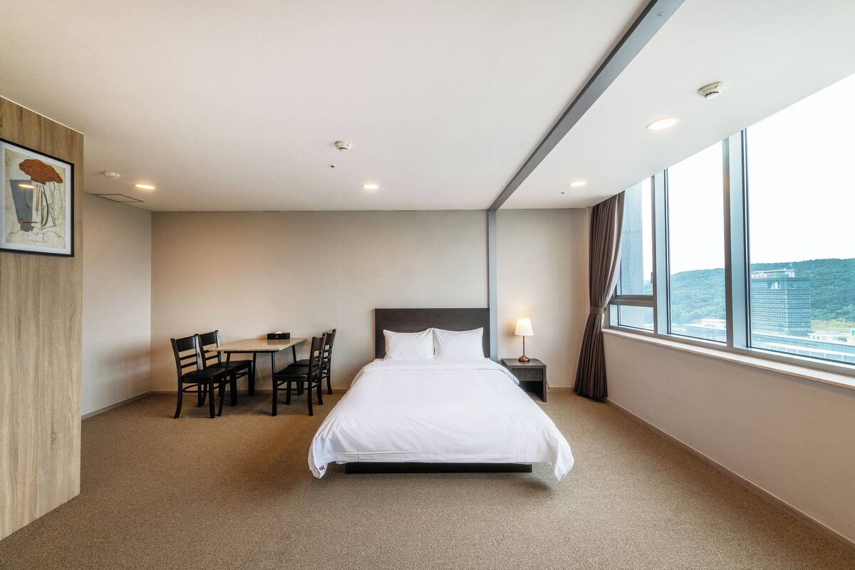 Une chambre d’hôtel avec un grand lit, un bureau avec des chaises, une fenêtre donnant sur un beau paysage et un tableau accroché au mur.