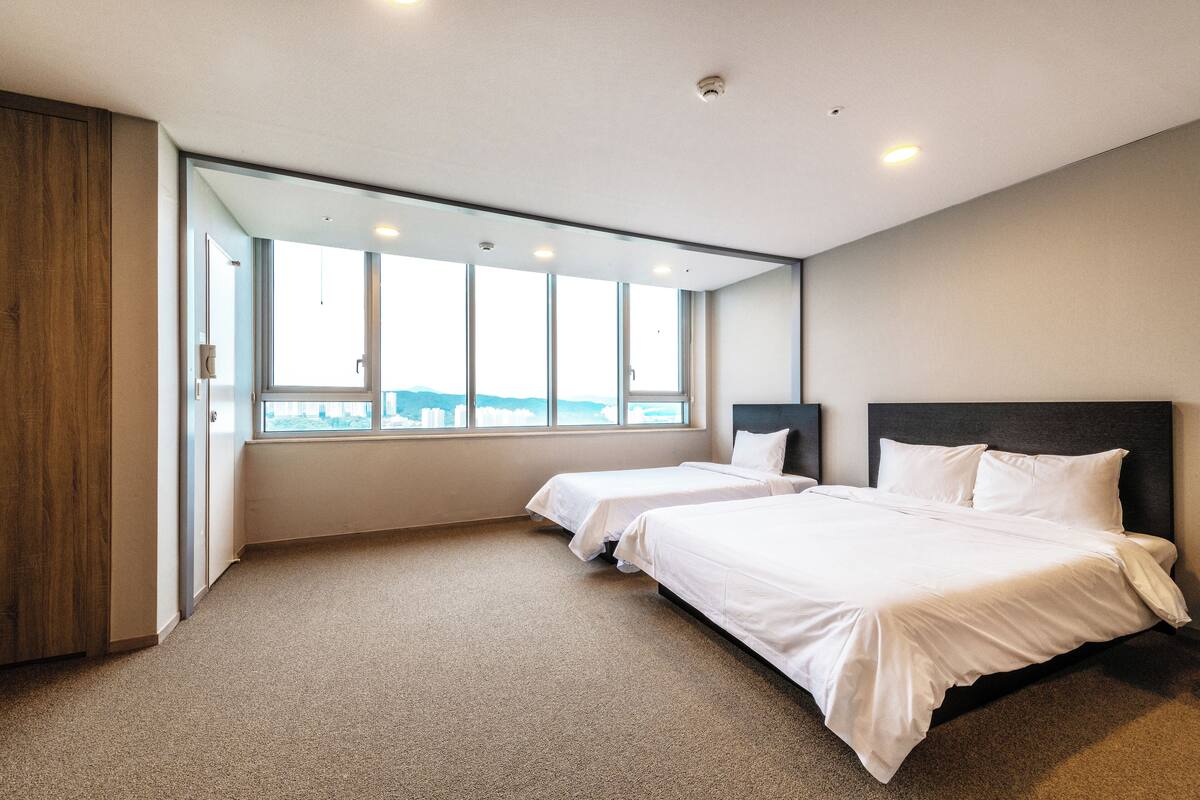 Une chambre d’hôtel moderne avec deux lits simples, une grande fenêtre donnant sur la ville et une armoire en bois.