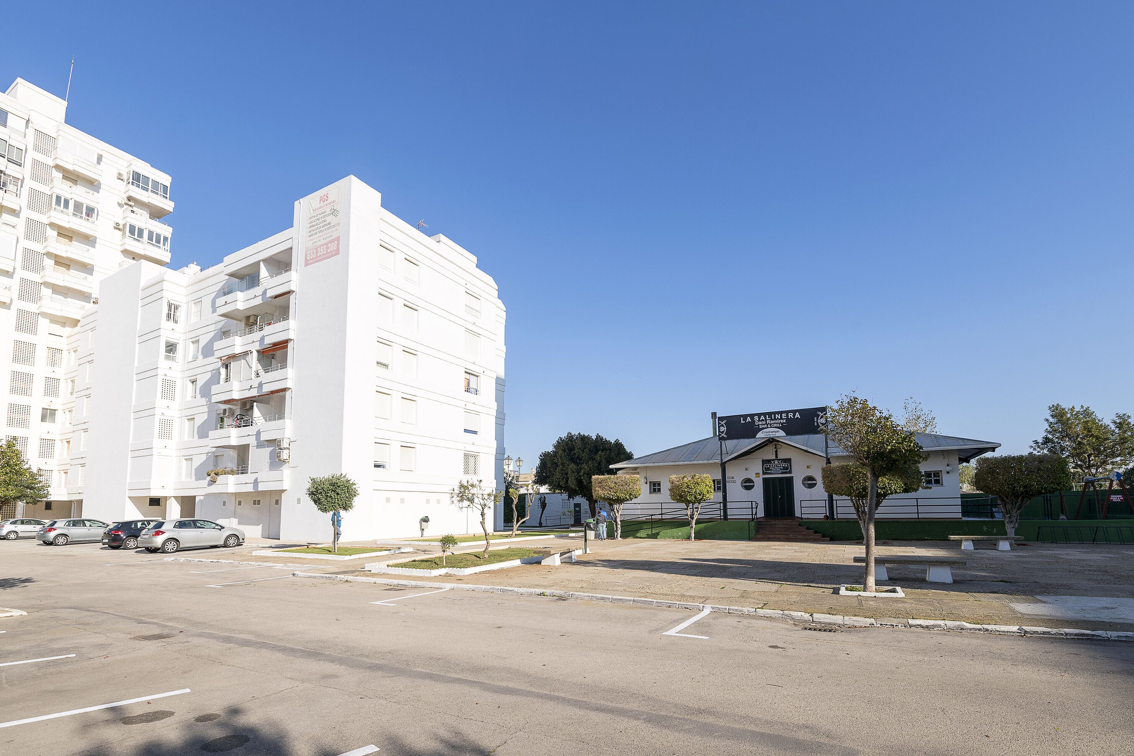 A multi-story white building with balconies, a parking lot, and a smaller building with a sign.