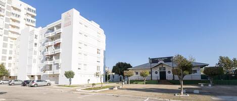 A multi-story white building with balconies, a parking lot, and a smaller building with a sign.
