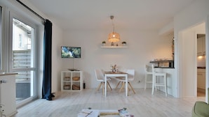 A modern living room with a glass coffee table, white furniture, a TV mounted on the wall, and a kitchen area visible through an open door.