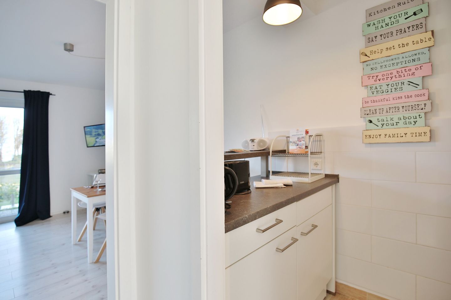 A modern kitchen with white cabinets, a wooden countertop, and a coffee machine. There are kitchen rules displayed on the wall.