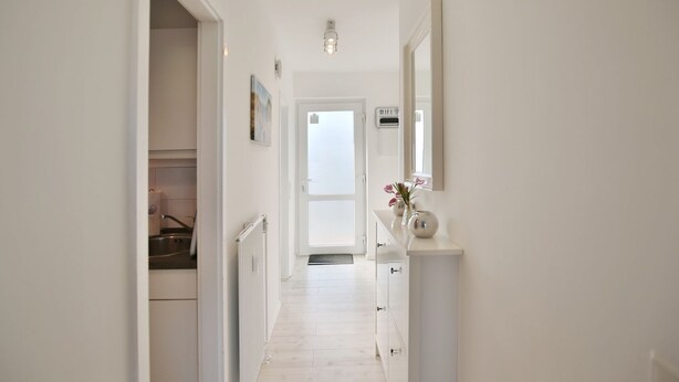 A narrow hallway with white walls, a radiator, and a door leading to a room. There is a small table with a vase of flowers and a mirror on the wall.