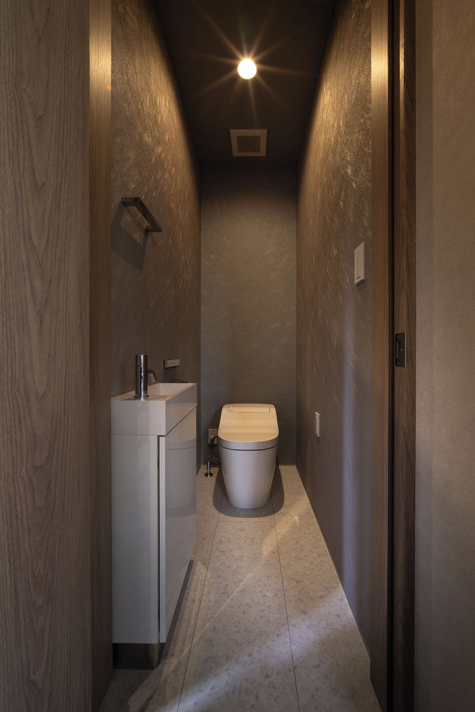 A narrow hallway with a modern bathroom featuring a toilet, sink, and a light fixture on the ceiling.