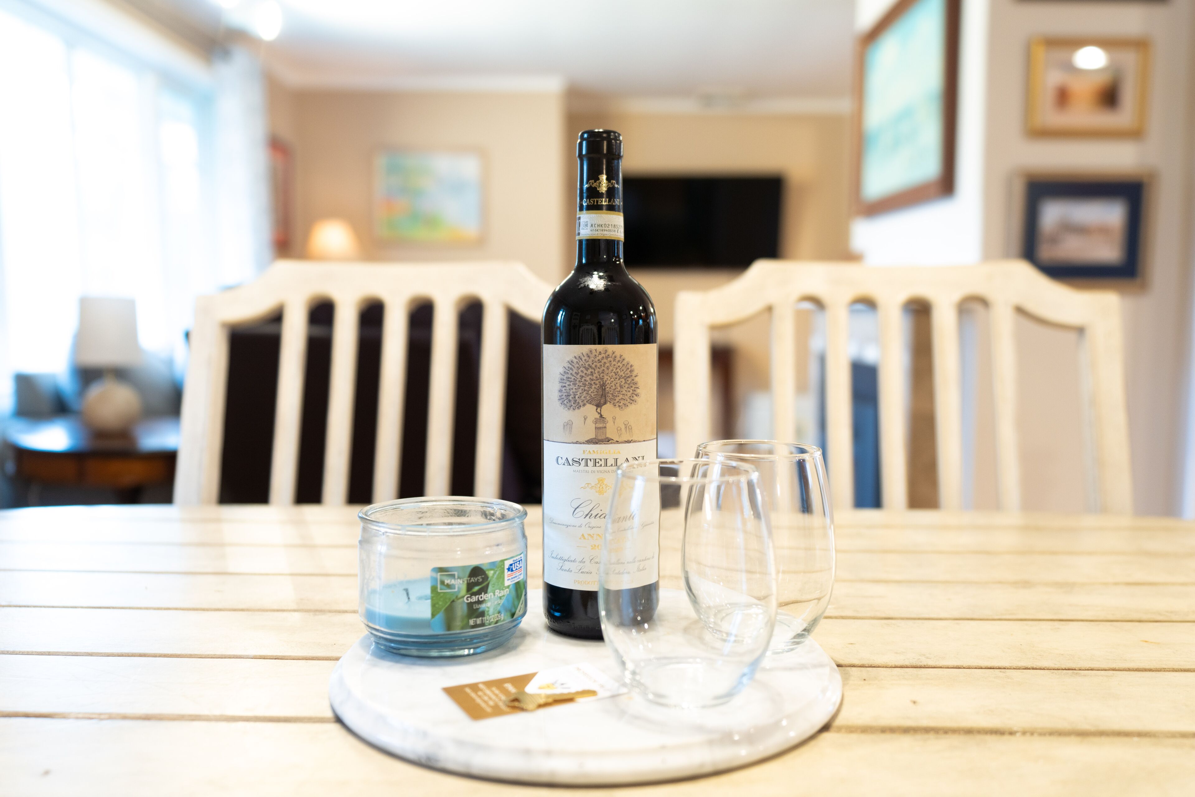 Una botella de vino, dos copas y una vela sobre una mesa en una habitación con sillas blancas y un televisor al fondo.
