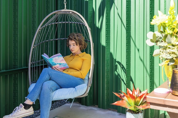 A person sitting in a hanging chair reading a book, with a green wall and a potted plant in the foreground.