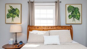 Een opgemaakt bed met patroonbeddengoed, een bedlampje, een klok en twee ingelijste botanische prints aan de muur.