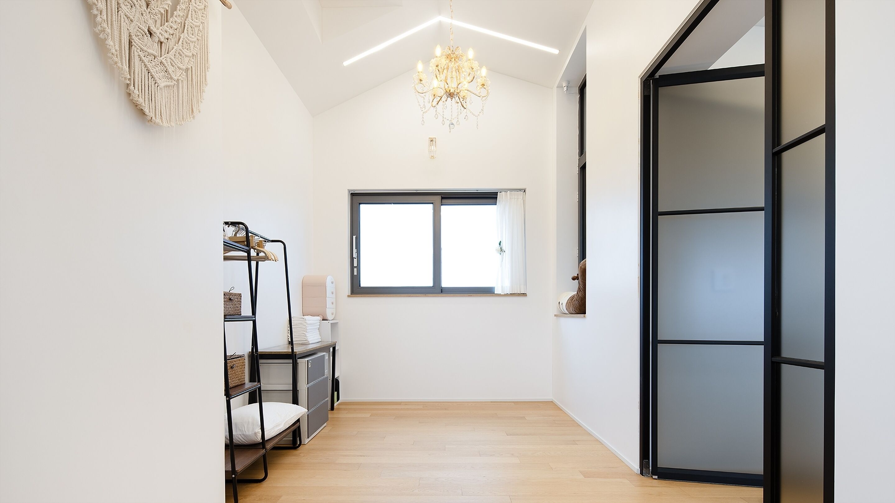 A hallway with a wooden floor, a black-framed door, a window with white curtains, a chandelier, and a macrame wall hanging.
