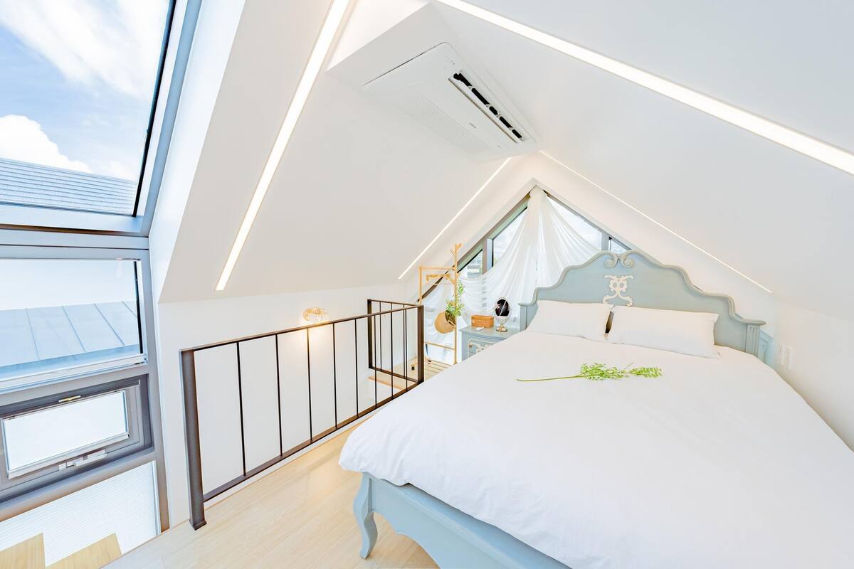 Un dormitorio con techo inclinado, una cama con ropa de cama blanca, una mesita de noche con una lámpara y una ventana con vistas al cielo.
