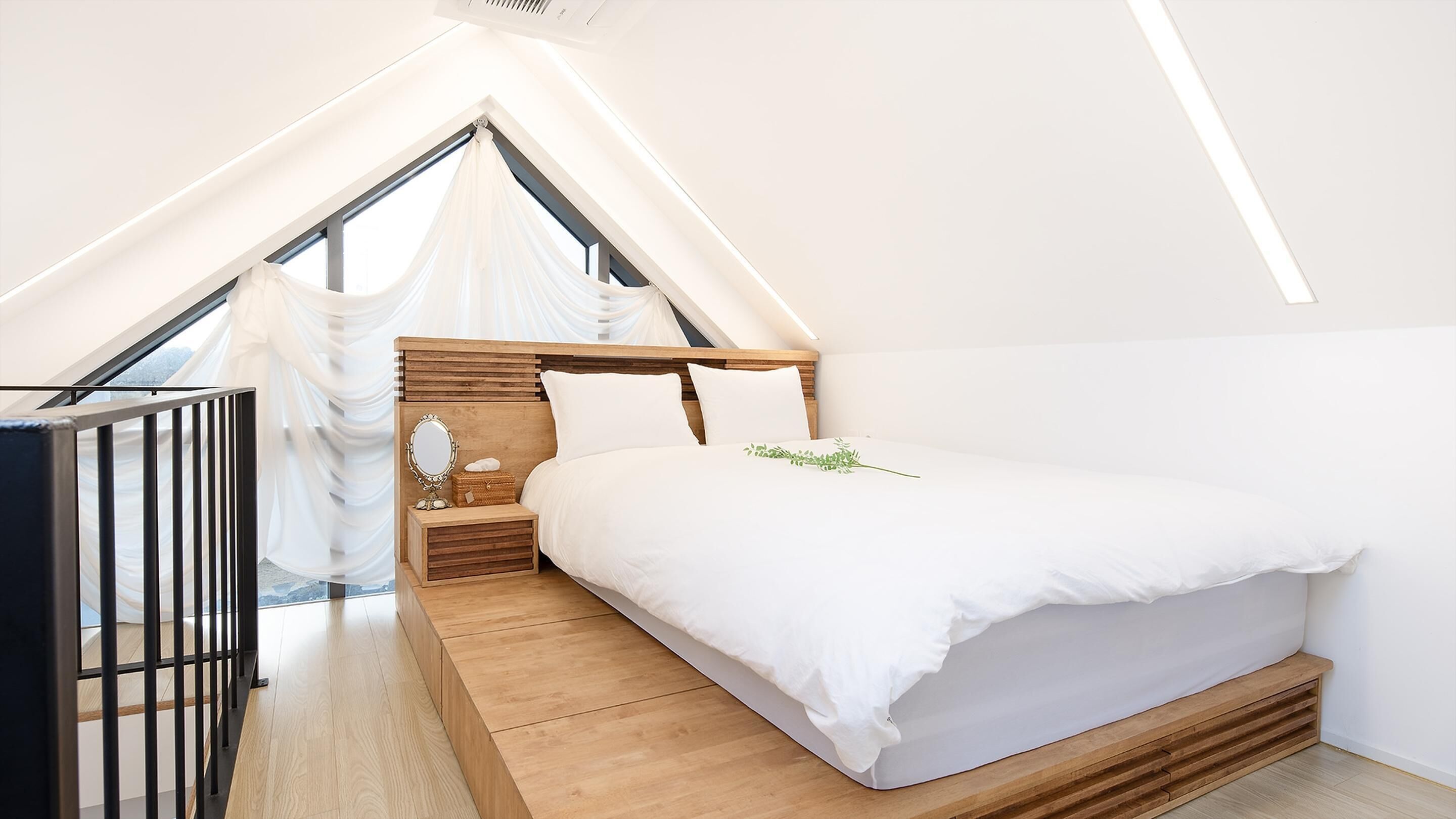A bedroom with a sloped ceiling, a wooden bed frame, a bedside table with a mirror, and a small window with white curtains.