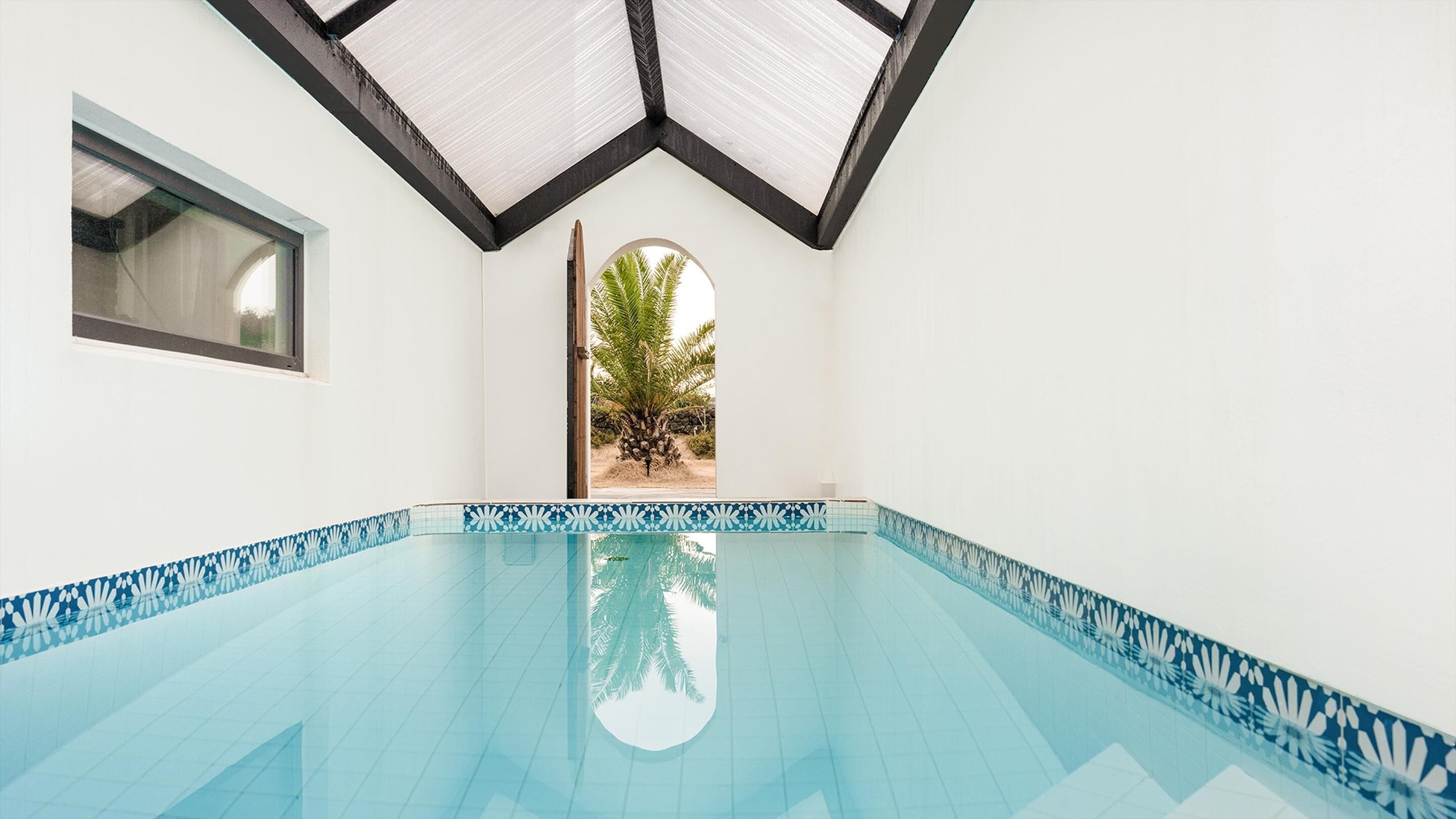 A swimming pool with a geometric ceiling and a view of a palm tree outside.