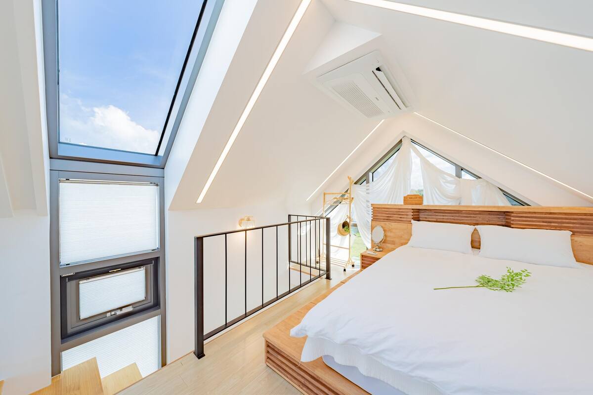 Un dormitorio moderno con tragaluz, cama con ropa de cama blanca, cabecero de madera y una mesita de noche pequeña con espejo.