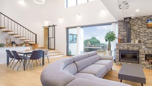 A modern living room with a grey sofa, a dining area with a table and chairs, a kitchen with wooden shelves, and a fireplace. - Jeju Geunalohu Pension (Jeju City)