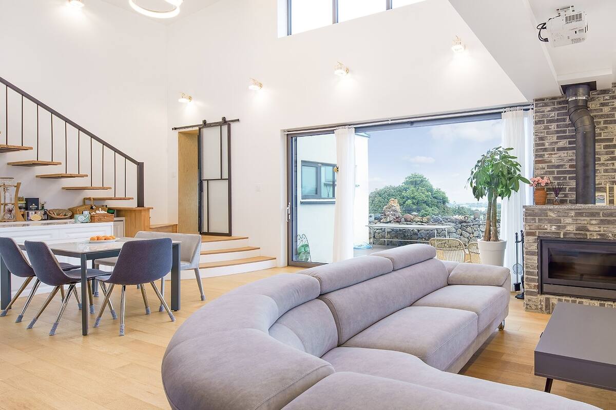 Una sala de estar moderna con un sofá gris, un comedor con mesa y sillas, una cocina con estanterías de madera y una chimenea.