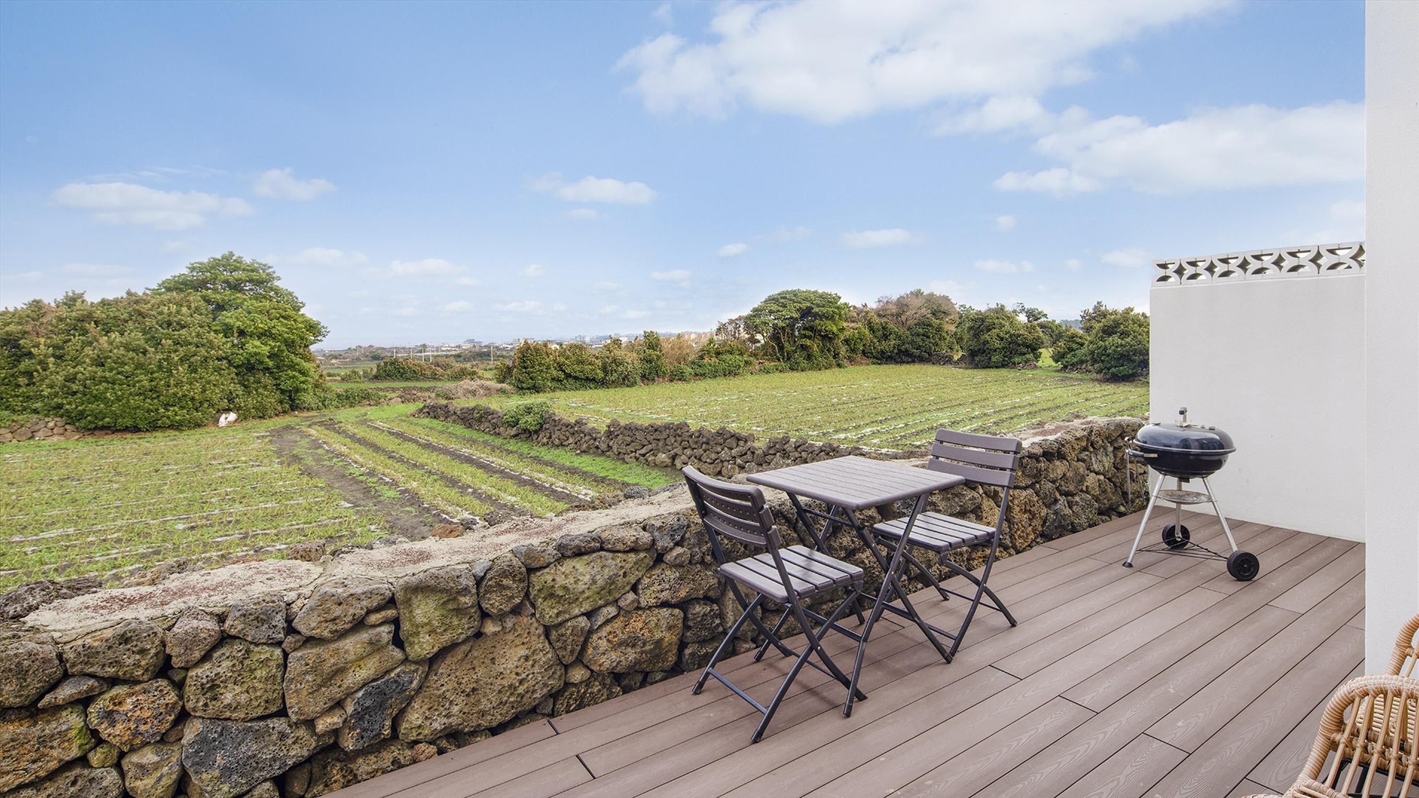 A patio with a stone wall, a table, chairs, and a barbecue grill overlooking a vineyard.