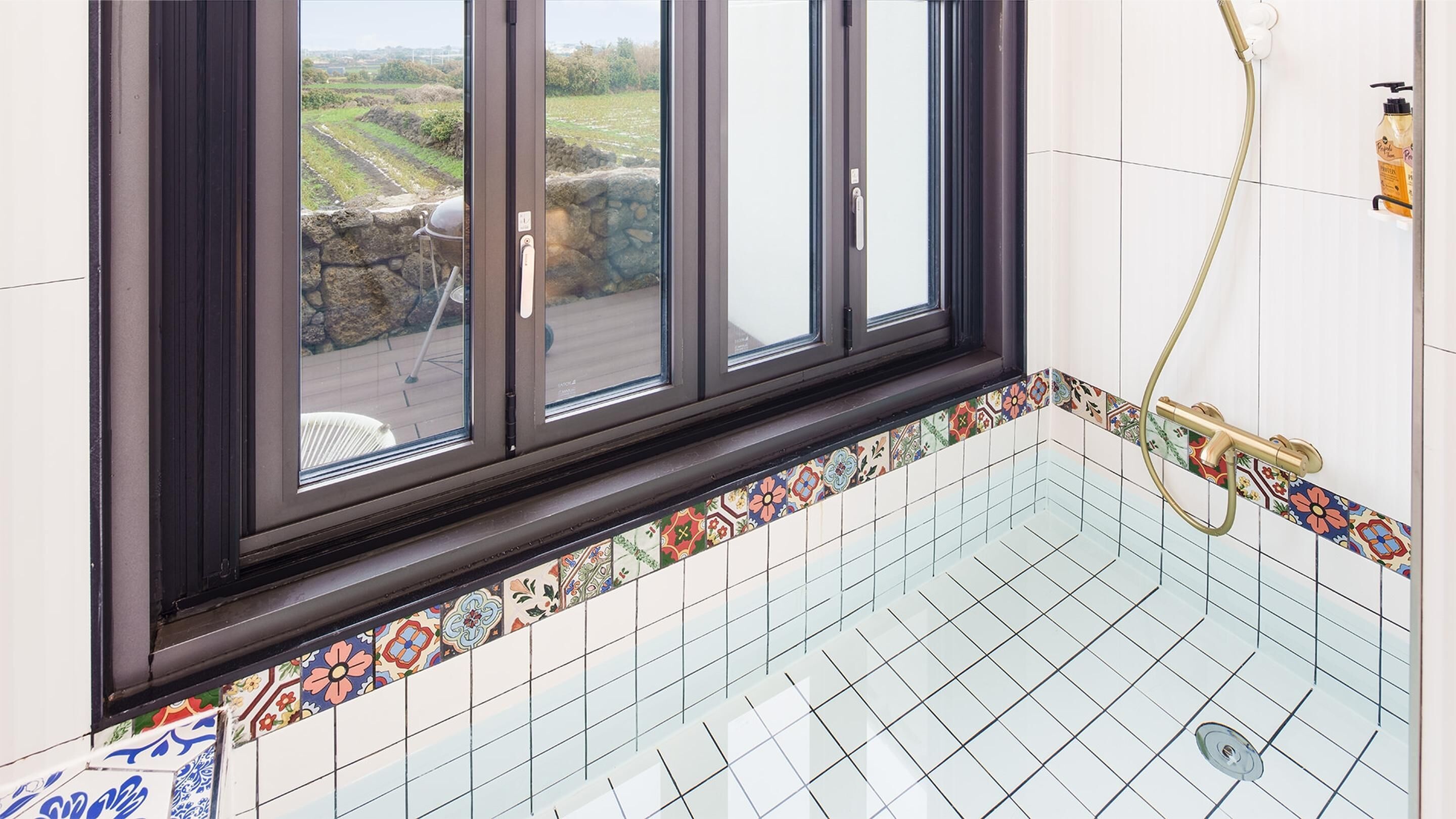 A bathroom with a shower, a window with a view of a garden, and a tiled floor.