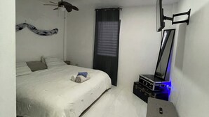 Un dormitorio con cama, ventilador de techo, televisor montado en la pared y un mueble pequeño.