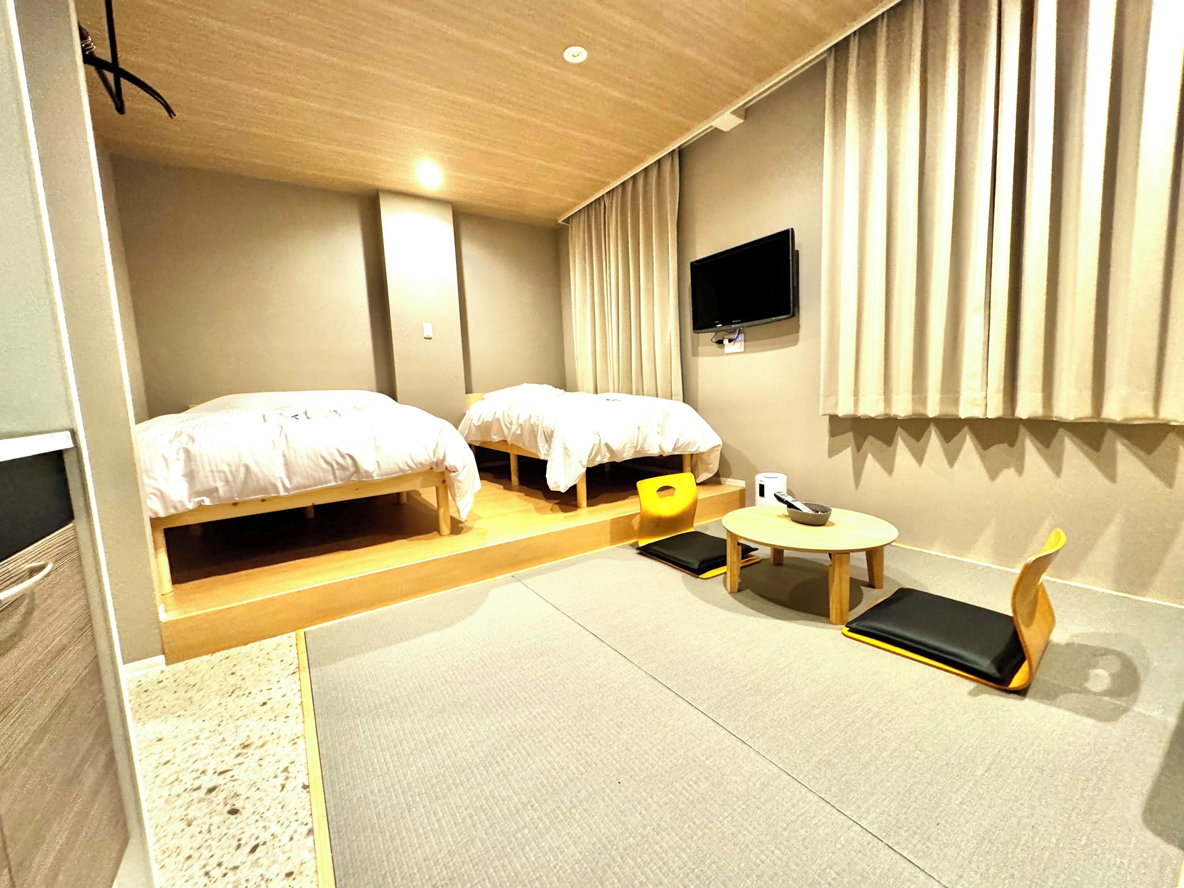 A hotel room with two beds, a small table, a chair, a TV, and a window with curtains.
